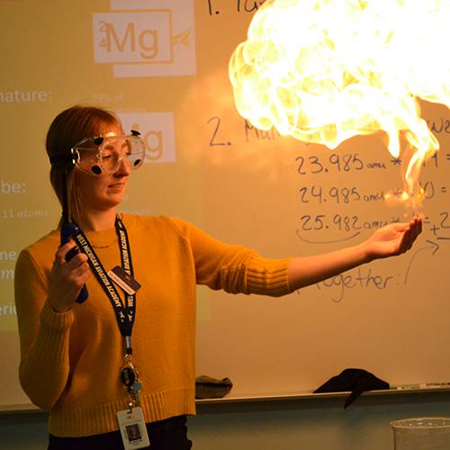 A teacher demonstrates a chemical reaction by creating a small fire ball above her outstretched hand